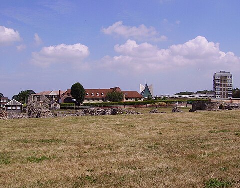 Landscape photo dominated by a field in front, surmounted by blue sky with white clouds. A row of modern buildings crosses the center; in front of the buildings a few stones can be seen piled atop each other.