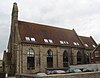 Side view of a tall, narrow flint building with a steeply-pitched red-tiled roof in which several skylights have been inserted at irregular intervals. Four identical lancet window openings with modern windows, separated by buttresses, dominate the near side; an identical window is visible on the recessed side wall behind it. A stone bell-tower rises from the left-hand side, and below it a porch projects forward a short way.