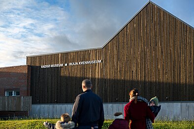 To voksne og tre barn foran vegg med tydelig logo der det står "Vadsø museum-Ruija kvenmuseum".