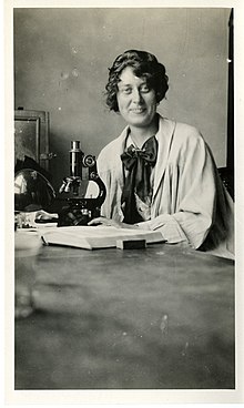 A smiling young white woman with dark curly hair, seated at a lab table, in front of a microscope