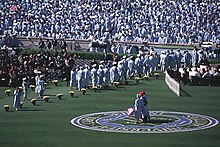 A bunch of people standing with cap and gowns while two people stand on a grass field.