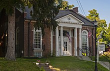 A small brick building in a neoclassical style, with two columns beside the main entrance and a gabled roof, seen from its front right, in the shade of a tall spruce tree. A sign next to the steps up from the street says "Hillsdale Town Hall".