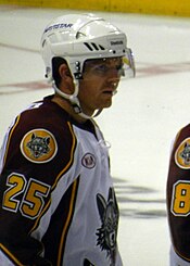 A Caucasian ice hockey player shown from the waist up. He wears a white helmet with a visor and a white jersey with maroon shoulders and sleeves. The number 25 is on his sleeve in yellow below a patch with the Wolves' logo.