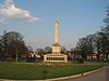 A curved area of grass with a floral border beyond which is a cream-coloured obelisk on a large plinth; in the distance are leafless trees and houses