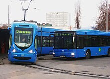 Zdjęcie przedstawia środki transportu miejskiego:tramwaj i autobus. Są niebieskiego koloru. Tramwaj linii 12 kieruję się w stronę Dubravy. Końcowym przystankiem autobusu linii 115 jest Skansko. W tle widać wysoki, biały budynek i drzewa.