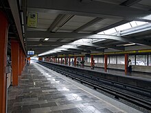 Picture of the Line 5 side platforms, where some people stands. The platforms are made of marble. In the far background, to the right, some cars are visible.