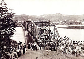 An old black and white photograph with a crowd on the end of embankment for a steel bridge which recedes into the hilly background spanning the river. On one side the bridge are tracks with a steam locomotive. On the other side is occupied horse drawn wagons.