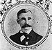 Head and shoulders of a white man with a wide bushy mustache, wearing a bow tie and dark suit with a medal pinned to the left breast. The portrait is surrounded by a circular frame and images of flowers.