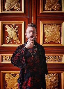 A half-length portrait of Canadian artist, Alexander McLeod (shown in a pensive pose, touching his chin), standing in front of a set of historical gilded, carved wood French doors during his visit to the Louvre in the spring of 2024. McLeod is wearing custom black and burgundy devore velvet ORXSTRA apparel, designed by Tala Kamea for presentation at Paris Fashion Week.