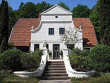 Frontale Farbfotografie von einem weißen Haus im Jugendstil, umringt von Bäumen und Büschen. Das hintere Hausteil hat ein rotes, tiefes Dach. Das angebaute Giebeldach hat eine Dreieckspitze und endet mit zwei Kurven, auf denen antik wirkende Vasen sind. Diese Vasen finden sich auch auf dem weißen Treppenabgang, der von der Terrasse in den Garten führt.