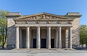 Farbfotografie eines klassizistischen Gebäudes mit sechs Säulen vor dem Eingang, über denen Figuren sind. Über den Figuren ist ein Giebelfeld mit Kampfszene.