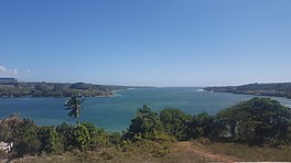 A photo taken from on top of a hill or cliff, overlooking a large bay. An entrance to the bay can be seen between flat landmasses on the right and left.