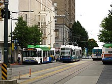 A streetcar and several short buses seen sharing a four-lane street with overhead wires and streetlamps.