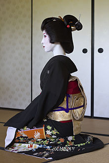 A profile view of a geisha sat kneeling. She wears a black formal kimono a gold belt, a traditionally styled wig and white make-up with red lips and accents.