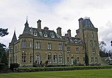 Ein imposantes zweistöckiges Haus mit gelben Steinen. Links befinden sich zwei Tourellen mit konischen Dächern, rechts steht ein größerer quadratischer Turm mit Pyramidendach. Auf dem Dach sind Dachgauben und Schornsteine sichtbar.