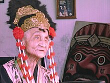 Portrait view of an elderly Indonesian woman wearing an elaborate headdress on the right, facing a painted traditional mask.