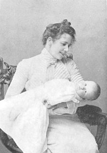 Black-and-white photo of Fohström holding a baby wrapped in a blanket. She smiles down at the baby while the baby looks at the camera.