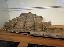 Wooden shipwreck remains displayed in a glass case on a wooden floor.