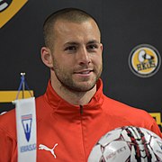 A man in a red jacket is sitting for a press conference behind a soccer ball.