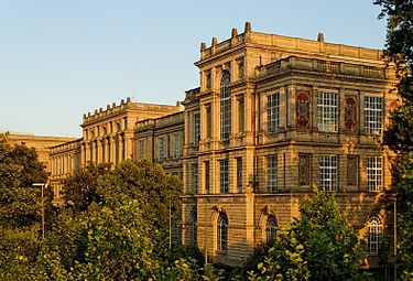 Seitliche Farbfotografie eines dreigeschossigen Gebäudes mit zwei Mittelteilen, die leicht vorgebaut und höher sind. Die hohen Fenster haben mehrere Sprossen und einige Rundbogenfenster befinden sich unter ihnen. Auf den Dächern stehen Steingeländer und Steinpfähle. An der rechten Seitenfassade sind zwischen den Fenstern hochkantige Reliefs. Das Gebäude wird von Bäumen umringt.