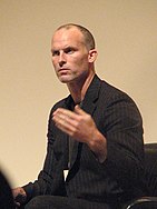 Matthew Barney sits and speaks. He has a light skin tone and a shaved head, and is wearing a black, striped suit and tie.