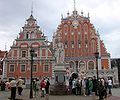 Frontale Farbfotografie von zwei weiß-braunen Gebäuden mit verzierten Staffelgiebeln. An jeder Staffelecke stehen Figuren und kleine Obelisken. An den Dachspitzen befinden sich Eisenkreuze. An der rechten Fassade hängen vier Statuen und vor dem rechten Gebäude ist ein flacher Vorbau mit einem verzierten Torbogen, in dessen Pfeilern zwei Farbfiguren stehen. Auf dem Vorplatz befindet sich eine weiße Ritterstatue mit Wappenschild, um die zwei Touristengruppen versammelt sind.