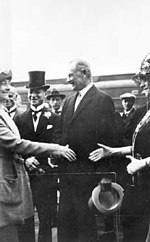 Two white woman extend their hands to one another, as if to shake them, while two middle-aged white men in formal attire look on.