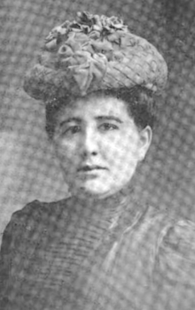 A woman dark hair and eyes, fair skin, wearing a hat and a dark dress with a high collar