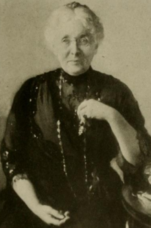 An older white woman, seated, with white hair parted center and dressed in an updo; she is wearing a black dress, and has one hand near her heart, and one hand in her lap