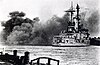 Bir savaş gemisi, ateş eden topların dumanları görünüyor.