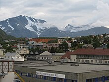 Foto einer Ortschaft, dahinter erhebt sich ein teils Schnee bedeckter Berg