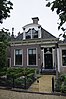 Dorpshuis met door pilasters geflankeerde ingang onder schilddak met grote versierde dakkapel en twee topschoorstenen met borden