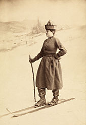 A young woman in a conical hat, with heavy clothing and boots with skis, stands on a shallow snow slope facing left. She holds a ski stick in her right hand.