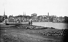 Wide view of a cityscape with evident destruction. Unused cannons and cannonballs litter the foreground, while a large Neoclassical building stands intact in the rear center.