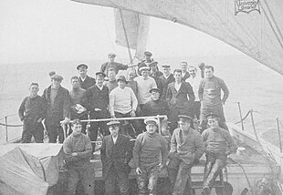 A group of men on board a ship, identified by a caption as "The Weddell Sea Party". They are dressed in various fashions, mostly with jerseys and peaked or other hats. The rough sea in the background suggests they are sailing into stormy weather.