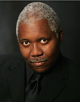 Head and shoulders of an older African American man with short, gray hair in all-black formal wear