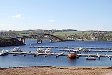 Foto einer Brücke über ein Gewässer. Auf der gegenüberliegenden Seite liegt eine Ortschaft mit einer Kirche. Vorne im Bild mehrere Boote an einer Marina.