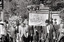A protest march, with a sign "No More Birminghams" prominent. Some of the marchers are black and some are white; all are well-dressed.
