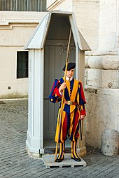 Frontale Farbfotografie von einem jungen Mann in blau-gelb-roter Uniform der Schweizergarde. Er trägt eine schwarze Baskenmütze und hält in der rechten Hand einen Speer. Er steht vor einem hellen Wachhäuschen aus Holz, das sich vor hellen Mauern eines großen Gebäudes befindet.