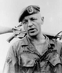 Head and shoulders of a white man wearing a t-shirt and a military jacket, unbuttoned at the collar, with the word "Rabel" on his right breast and "U.S. Army" on his left. He has on a beret and is holding a rifle over his shoulder by its barrel.