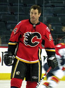 A man with short, brown hair looks to his right. He is wearing a red uniform with black pants and a black stylized C logo on his chest.