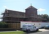 A long brick and tile building with central tower on the near side. The whole structure is surrounded by scaffolding and barricades. The tower has tall round-headed windows and louvres on each face, a shallow tiled cap and a stone crucifix.