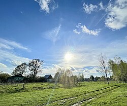 A rural russian village is depicted. In the foreground, we see a green field and behind it a road with a row of houses.