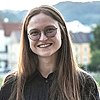 Alina Gorlova schaut lächelnd in die Kamera, sie trägt ihr braunes mehr als schulterlanges Haar mittig gescheitelt offen. Ihre Brille ist rund und ihr Oberteil schwarz.