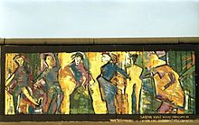 Frontale Farbfotografie von der Berliner Mauer mit einem Bild im expressionistischen Stil. Sechs Menschen bewegen sich auf gelb-grünem Untergrund und unten rechts steht „Sabine Kunz Halle Mühlweg 29“.