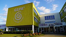 Firmengebäude der GEPA. Es zeigt das GEPA-Logo in grau auf grünem Grund. Fotos von Mitarbeitenden sowie Handelspartnern sind außen ebenfalls angebracht.