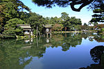 A stone lantern next to a tree in front of a pond. Beyond the lake there is a small wooden house built on poles above the water.