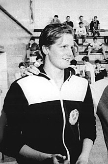 Swimmer Kristin Otto shown from the waist up wearing a jacket, with spectators on bleachers that can be seen in the background