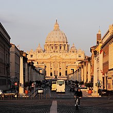 Frontale Farbfotografie von der Hauptstraße mit Blick auf den Petersdom, der von der untergehenden Sonne angestrahlt wird. Der dreigeschossige Dom im Renaissance-Stil hat zwei kleine Kuppeln und eine große Kuppel mit Kreuz. Entlang der Straße führen Säulen mit Laternen.