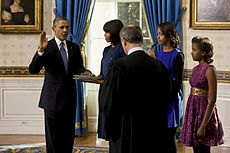 Barack Obama holds his right hand in the air as Michelle Obama looks at him and Malia and Sasha Obama watch as a balding man whose back is to the camera looks down.